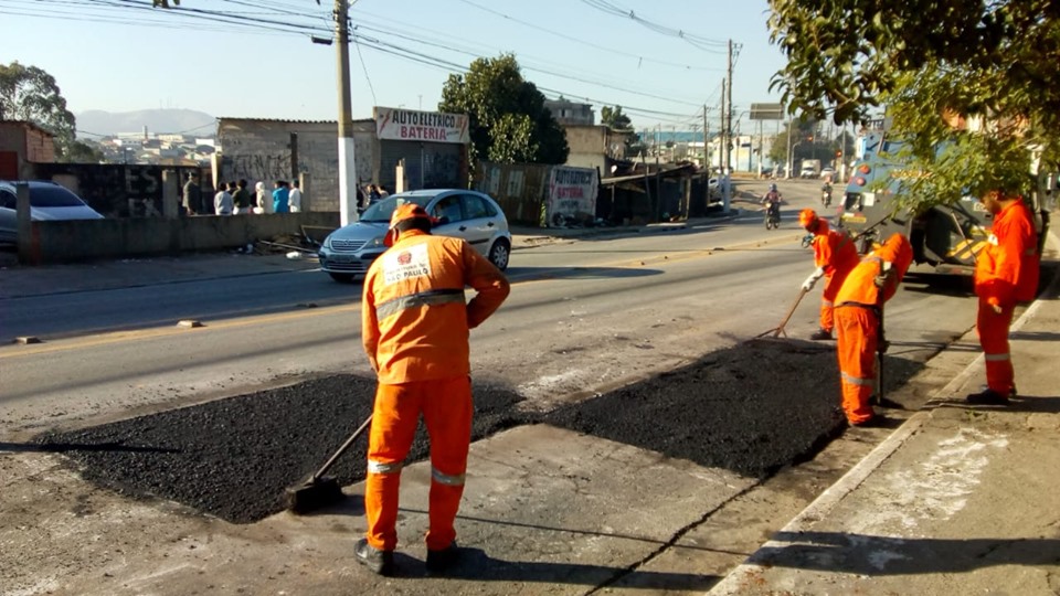 Funcionários nivelando o asfalto utilizado para tapar os buracos na rua Morro das Pedras, sob sol.